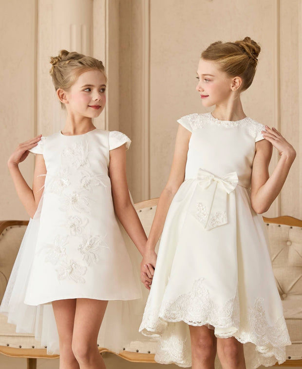 Two young girls wearing white dresses holding hands in a room with beige walls. 