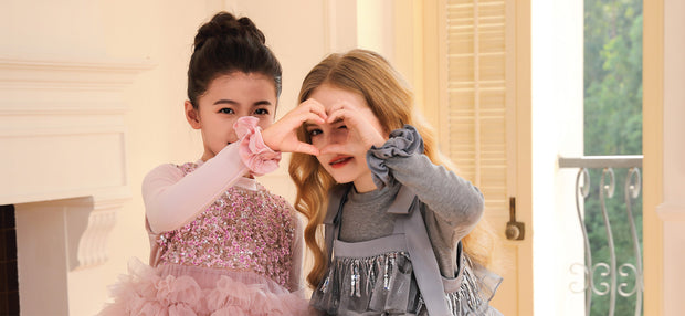 Girls in festive Tulleen holiday dresses make a heart shape with their hands, radiating seasonal joy