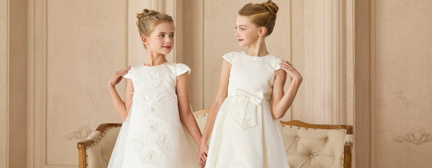 Two young girls in white dresses standing in a room with beige walls and furniture.