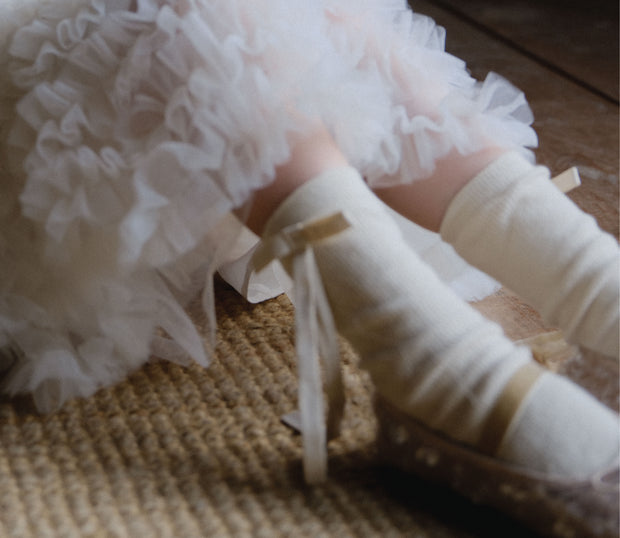 Girl in white Tulleen tutu skirt and ribboned socks, seated on woven rug in soft ballet light.